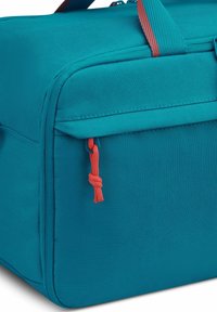 Bolsa de tela en color azul verdoso con forma rectangular, tirador de cremallera rojo que contrasta y superficie texturizada. Presenta un bolsillo lateral con un cordón de acento.