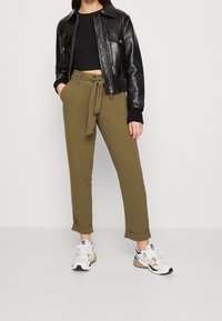 Zwarte cropped jas van nepleer met een ritssluiting, olijfgroene high-waisted broek met een gebonden taille, en witte, zwarte en beige sneakers.