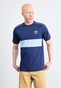 Tamno plava pamučna majica s svijetloplavom horizontalnom prugom, Adidas logotip na prsima, i zakrivljenim izrezom. Kratki rukavi s tri pruge na ramenima.
