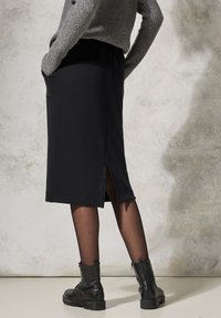Schwarze midi Länge Rock mit seitlichem Schlitz, elastischem Bund und Taschen. Gestylt mit einem grauen Pullover und schwarzen Knöchelstiefeln. Glatte Stoffbeschaffenheit.