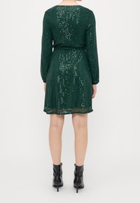 Femme portant une robe longue à manches longues en sequins vert foncé et des bottines noires à talons, vue de dos sur un fond uni.