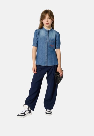 Jeune fille portant une chemise en jean à manches courtes, un pantalon bleu marine, des baskets noires et blanches, tenant une pochette noire à clous sur un fond uni.