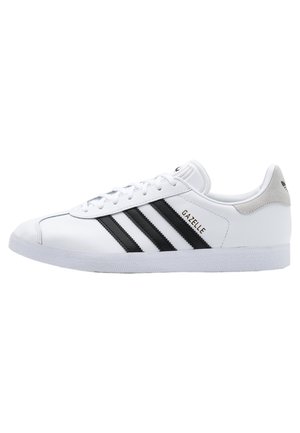 Biały niski sneaker Adidas Gazelle z czarnymi paskami, białymi sznurówkami i szarą zakładką na pięcie, pokazany w prawym profilu.
