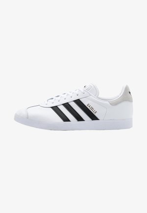 Zapatilla blanca Adidas Gazelle de caña baja con rayas negras, cordones blancos y lengüeta gris en el talón, mostrada en perfil derecho.