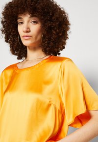 Blusa arancione in satin con scollatura rotonda e maniche corte e larghe. Il tessuto ha una texture liscia e riflettente. Nessun motivo o dettaglio prominente.