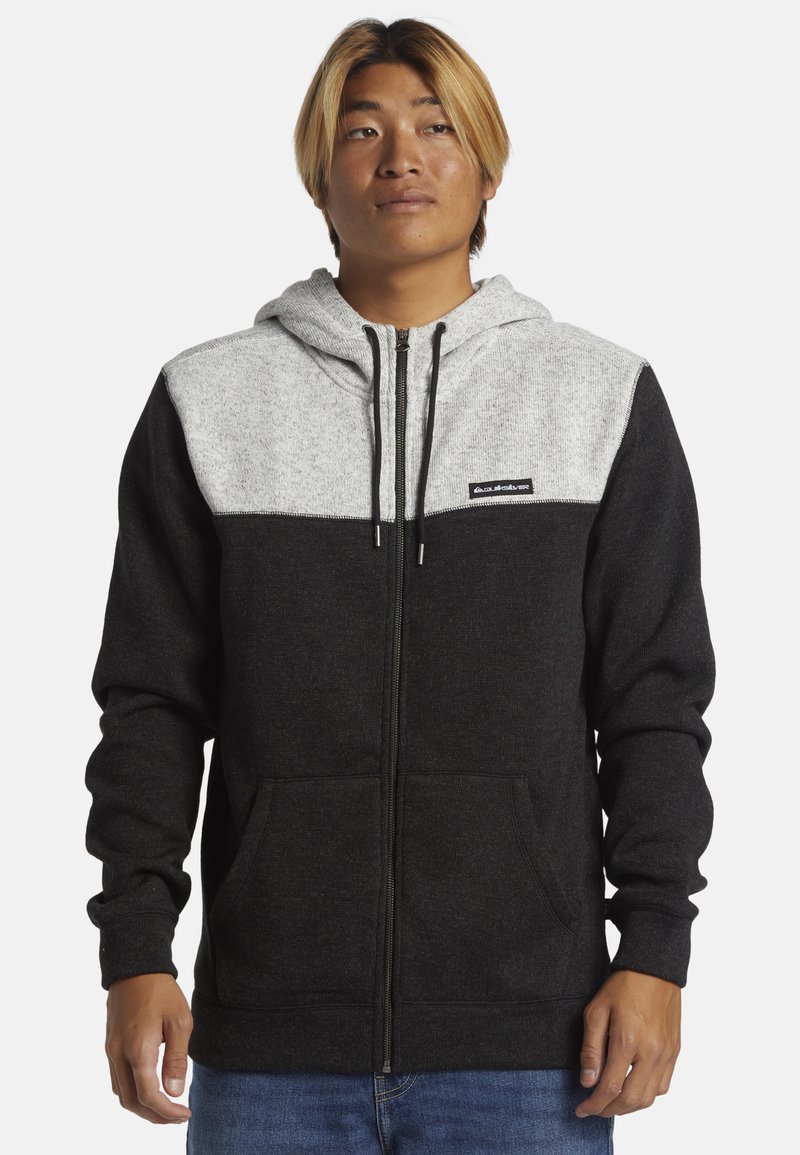 Haut à capuche gris et noir zippé en tissu doux, avec une capuche, des poches avant et une patch logo sur la poitrine.