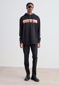 Czarna bluza z kapturem z różowym paskiem z logo Calvin Klein, w połączeniu z czarnymi spodniami i czarnymi butami; model noszący okulary przeciwsłoneczne stoi na neutralnym tle.