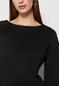 Haut noir à manches longues avec un large décolleté bateau. Le tissu semble lisse avec un subtile éclat et une coupe structurée.