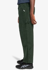 Groene corduroy broek met een ontspannen pasvorm, voorzien van zijzakken en een vlakke voorkant. Gepaard met zwarte sportieve schoenen.