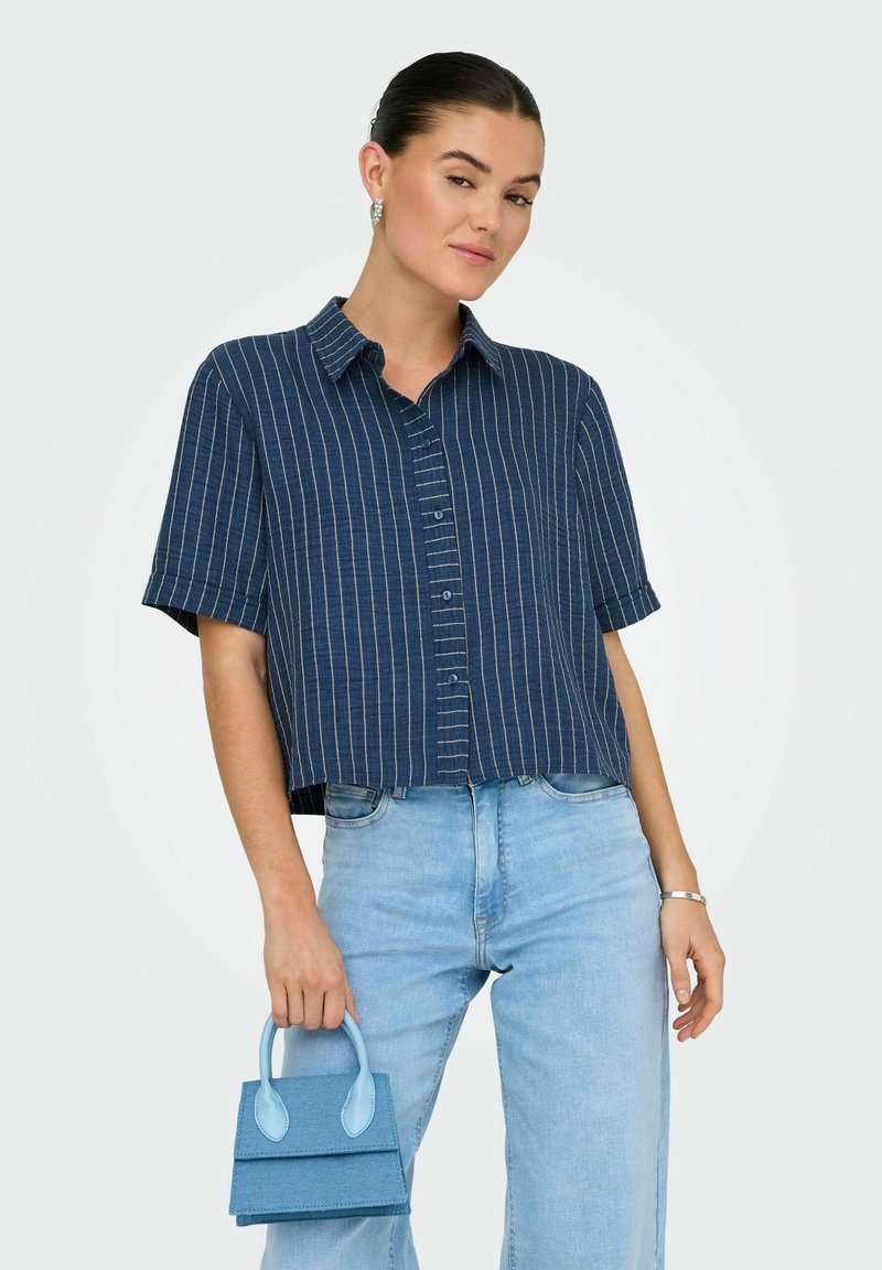 Chemise navy raccourcie avec des rayures verticales blanches, à boutons et à manches courtes. Associée à un pantalon en denim bleu clair et un petit sac à main bleu.