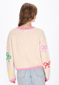 Cremfarbener Pullover mit rosa Akzenten, der bunte Schleifenapplikationen in Grün, Rot, Lila und Gelb zeigt. Hergestellt aus einem weichen, strukturierten Strickmaterial.