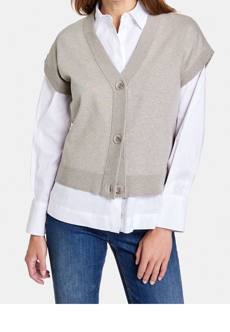 Gilet tricoté gris avec un col en V, des manches courtes et cinq boutons, superposé à une chemise blanche. Le tissu a une texture lisse.