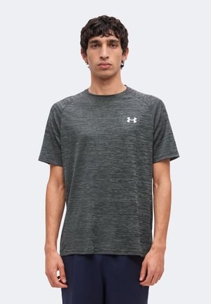 Jonge man met krullend donker haar draagt een grijs Under Armour T-shirt met korte mouwen en een donkere broek, staand tegen een eenvoudige lichte achtergrond.