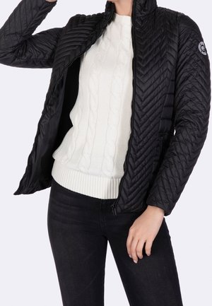 Giacca nera trapuntata con colletto alto e chiusura con zip, con una fantasia testurizzata. Indossata sopra un maglione in maglia a treccia color crema e jeans scuri.