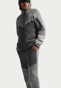 Mand iført mørkegrå og lysegrå Nike træningsdragt med hætte trukket op og hænder i lommerne, solbriller, stående mod en ensfarvet baggrund.