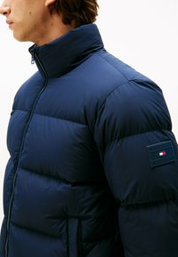Doudoune bleu marine avec un col haut, présentant des motifs de piquage horizontaux, des poches latérales et une patch de marque sur la manche.
