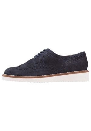 Marineblauwe suède brogue schoen met wingtip-stijl, perforaties, witte rubberen zool en een bruine rand rondom de zoolrand.