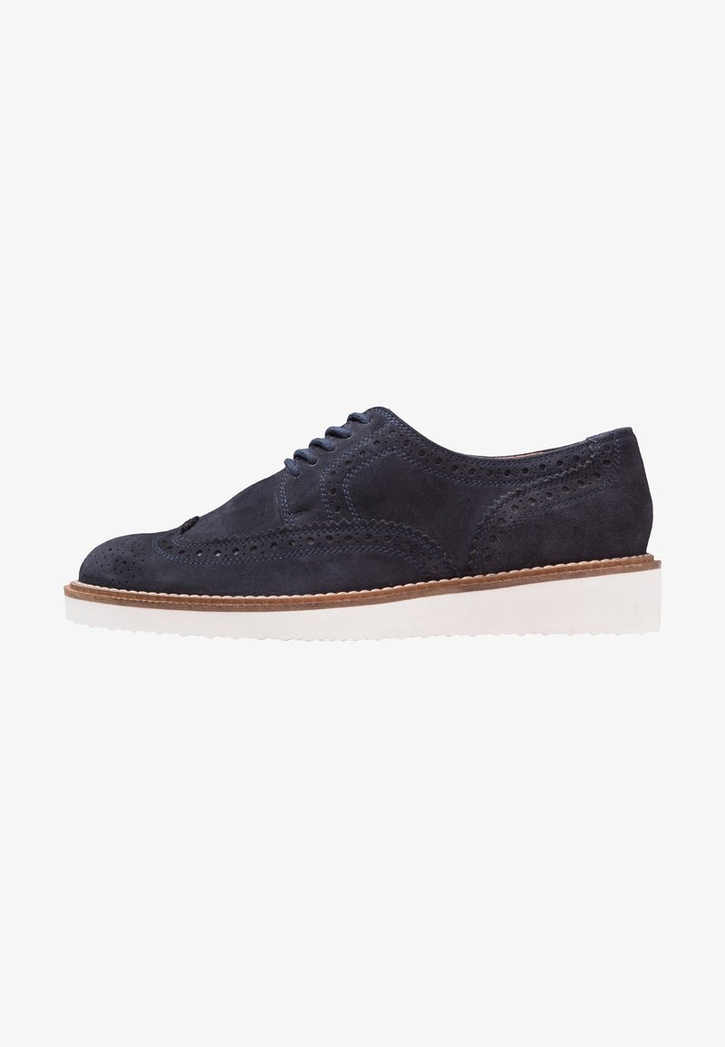 Chaussure brogue en daim bleu marine avec motif à ailettes, perforations, semelle en caoutchouc blanche et bordure brune autour du bord de la semelle.