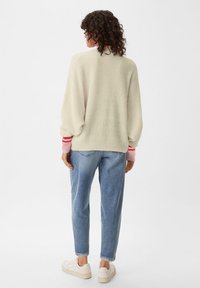 Krémový oversize pletený svetr s růžovými a červenými pruhovanými manžetami, v kombinaci se světle modrými zúženými džíny a bílými teniskami. Měkká textura a uvolněný střih.