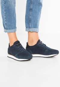 Stopy w granatowych zamszowych trampkach z białymi podeszwami i dopasowanymi sznurowadłami, w zestawieniu z podwiniętymi jasnoniebieskimi dżinsami na białej podłodze.