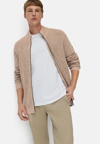 Strickjacke in Hellbraun mit Reißverschluss vorne, geripptem Kragen und langen Ärmeln, getragen über einem weißen T-Shirt, kombiniert mit beigen Hosen.