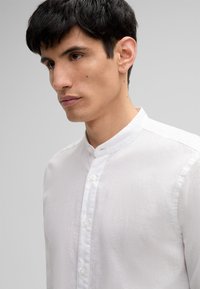 Giovane uomo con capelli neri corti che indossa una camicia bianca con colletto alla mandarina, che guarda leggermente verso il basso e a sinistra.