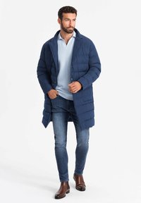 Abrigo acolchado azul marino con cuello de muesca, combinado con una camiseta polo azul clara y jeans de mezclilla. Botas marrones hasta el tobillo completan el look.