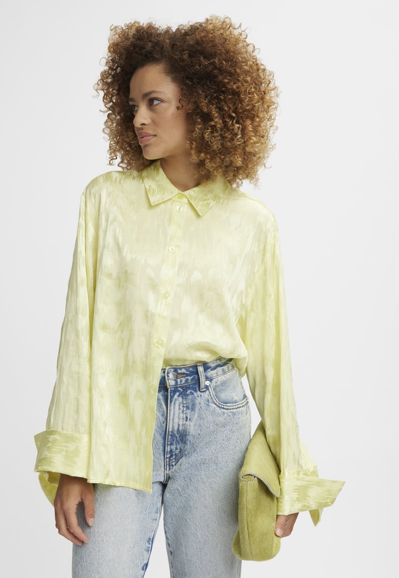 Blusa di raso giallo chiaro con bottoni frontali, maniche lunghe e una texture sottile, abbinata a jeans a vita alta con effetto acidato e una pochette verde.