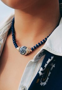 Collier de perles bleues avec une perle centrale en céramique blanche ornée d'un motif floral, des accents dorés et des perles cylindriques bleu foncé.