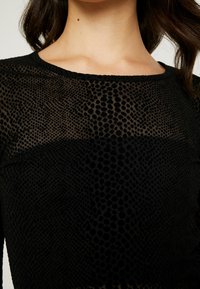 Svart topp med ett texturerat mesh-mönster med cirkulära detaljer. Halsringningen är rundad och tyget är lätt och genomskinligt.