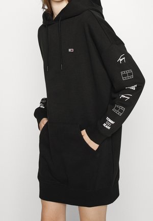 Person wearing oversized black hoodie dress with Tommy Jeans logos on chest and multiple white logos and text on right sleeve.