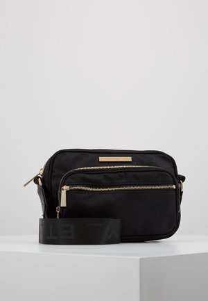 Zwarte nylon crossbodytas met gouden ritsen en verstelbare brede band, geplaatst op een wit oppervlak tegen een lichtgrijze achtergrond.