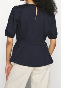 Marineblå peplum-top med korte puffærmer, en nøglehulåbning i ryggen og samlet taljedetalje. Lavet af et glat, let stof.