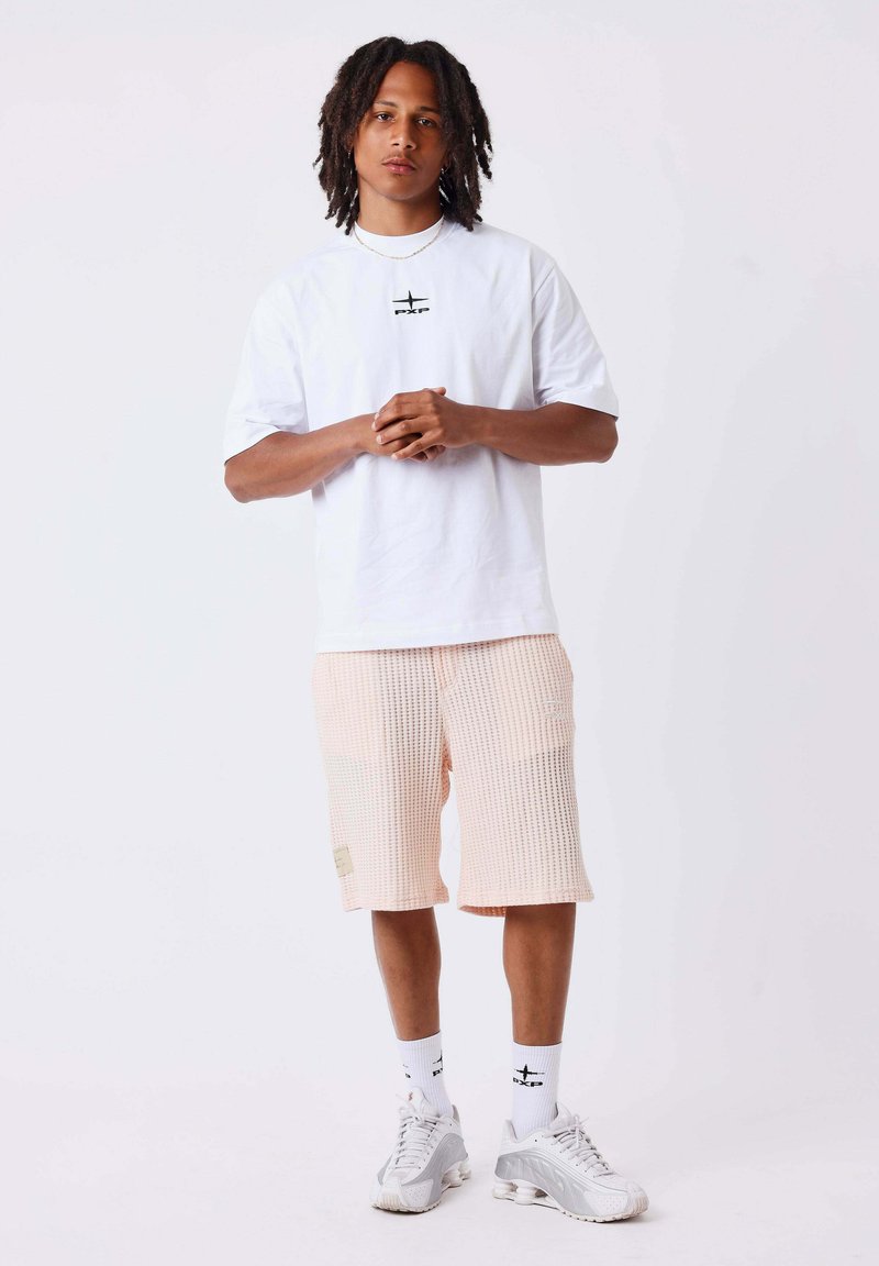 Homme avec dreadlocks portant un t-shirt blanc PXG, un short rose clair texturé, des chaussettes blanches et des baskets blanches sur un fond uni.