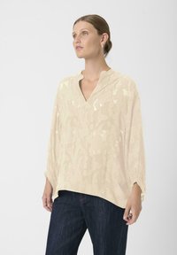 Šviesiai smėlio spalvos bliuska su tekstūriniu raštu, V-formos iškirpte ir ilgomis rankovėmis. Derinama su tamsiai mėlynomis džinsinėmis kelnėmis.