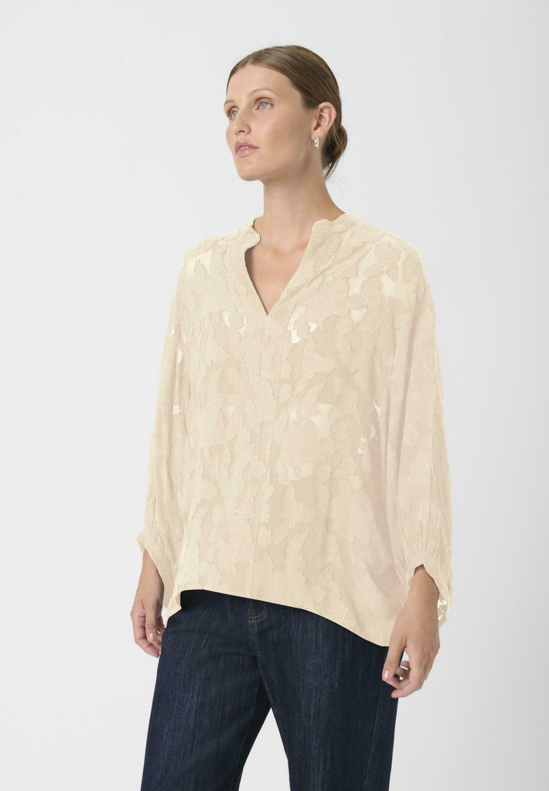 Šviesiai smėlio spalvos bliuska su tekstūriniu raštu, V-formos iškirpte ir ilgomis rankovėmis. Derinama su tamsiai mėlynomis džinsinėmis kelnėmis.