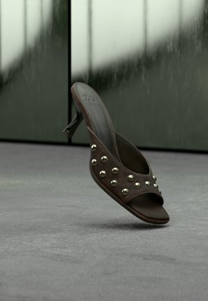 Mule à talons hauts marron foncé avec des clous dorés sur la bride, flottant au-dessus d'un sol gris devant des panneaux verdâtres flous.