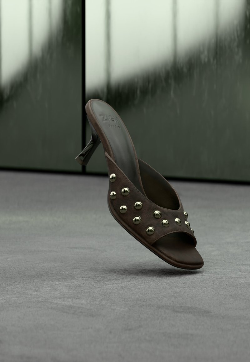 Σκούρο καφέ mule με ψηλό τακούνι και χρυσά καρφιά στο λουράκι, απεικονίζεται να αιωρείται πάνω από γκρι δάπεδο μπροστά από θολά πράσινα πάνελ.