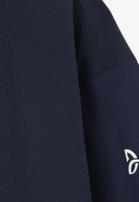 Prenda de color azul marino con textura suave, que presenta un sutil detalle del logo en blanco. El diseño incluye una zona de hombros estructurada.