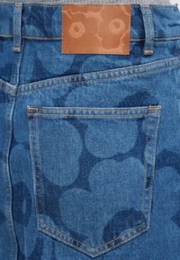 Farmerrövidnadrág zsebe sötét virágmintákkal és barna bőrpárnával, amely a derékpánton van varrva.