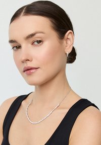 Kobieta o ciemnych włosach upiętych w kok, ubrana w czarną bezrękawnik, srebrny naszyjnik z małymi kamieniami i srebrne kolczyki w kształcie kółek na białym tle.