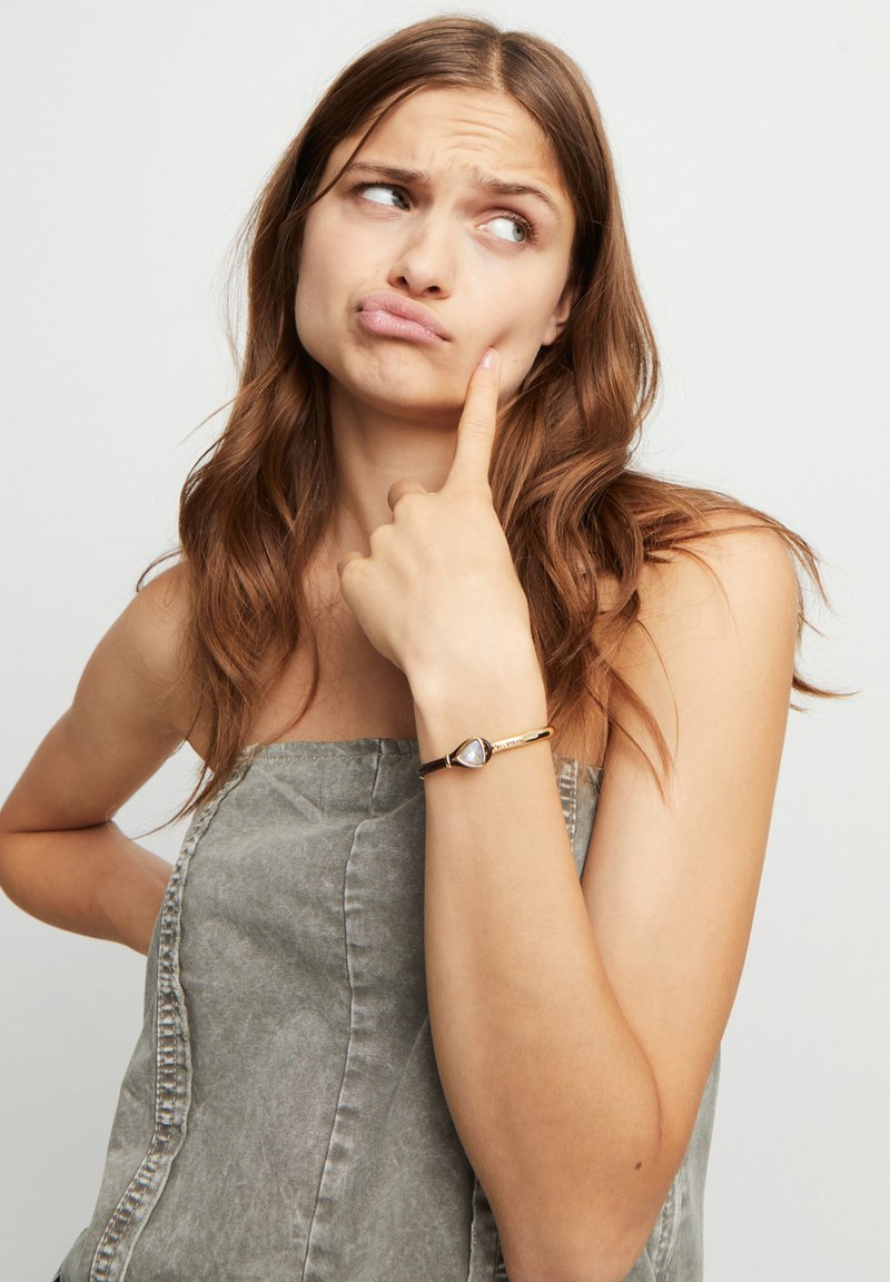 Bracelet en or avec une pierre précieuse en forme de goutte. Le modèle porte un haut sans manches gris, avec des cheveux mi-longs et une expression pensive.