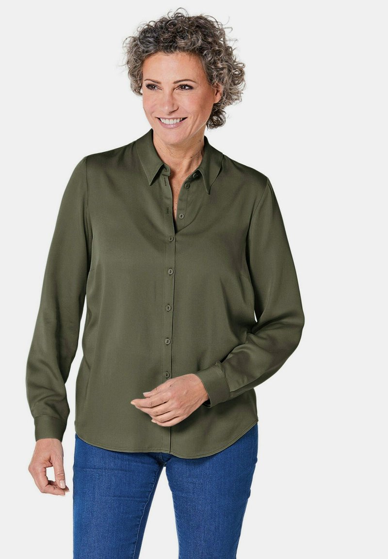Dunkelgrüne, langärmelige Button-Down-Bluse mit Kragen, die eine glatte Textur und einen abgerundeten Saum aufweist, kombiniert mit blauen Jeans.