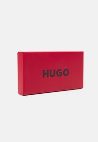 Röd rektangulär låda med matt yta, med den svarta texten "HUGO" tydligt synlig på framsidan. Inga synliga detaljer eller mönster.