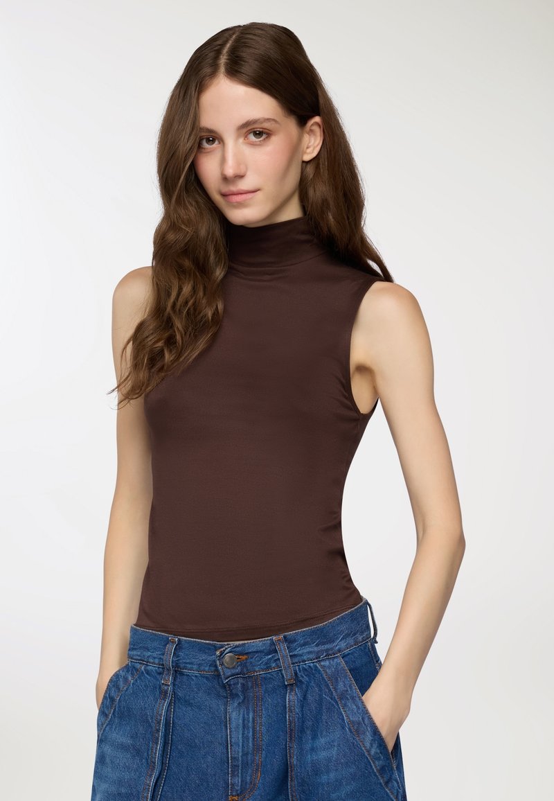 Brązowy bezrękawnik z kołnierzem w gładkim, elastycznym materiale, zestawiony z niebieskimi spodniami dżinsowymi z wysokim stanem i przednimi kieszeniami.