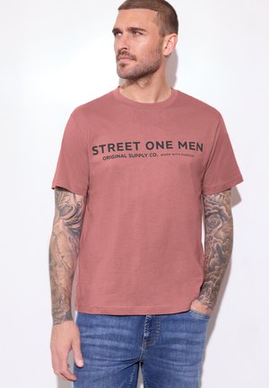 Mężczyzna o krótkich włosach i tatuażach na rękawach, ubrany w różową bluzkę z napisem „Street One Men” i niebieskie jeansy, patrzący na bok.