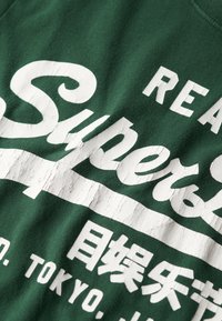Camiseta verde con texto blanco audaz y desgastado. La tela muestra una textura suave con un cuello redondo clásico.