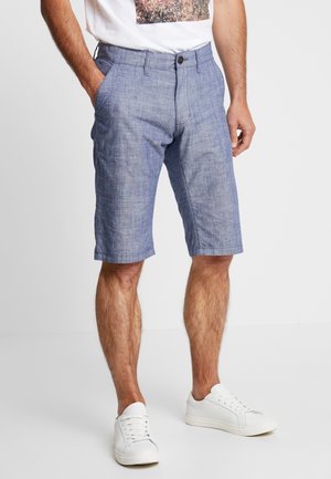 Homme portant un short bleu clair jusqu'aux genoux, des baskets blanches et un t-shirt blanc avec un imprimé graphique, debout avec une main dans la poche.