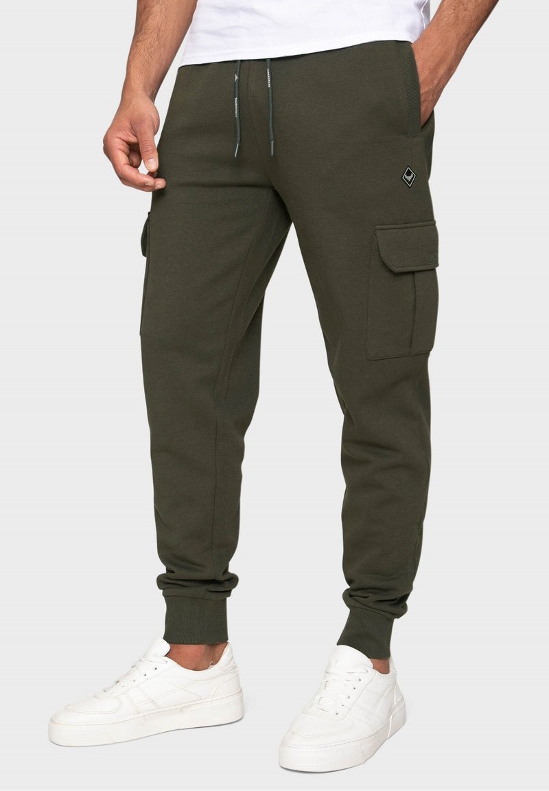 Pantalones de chándal verde oliva con puños elásticos y dos bolsillos laterales tipo cargo, hechos de una tela suave, con un logo decorativo en la parte frontal.
