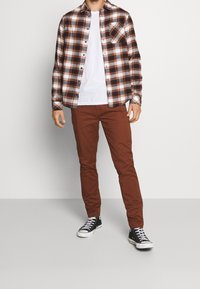 Un homme portant une chemise à carreaux marron par-dessus un t-shirt blanc, un pantalon couleur rouille et des baskets montantes noires, se tenant devant un fond uni.